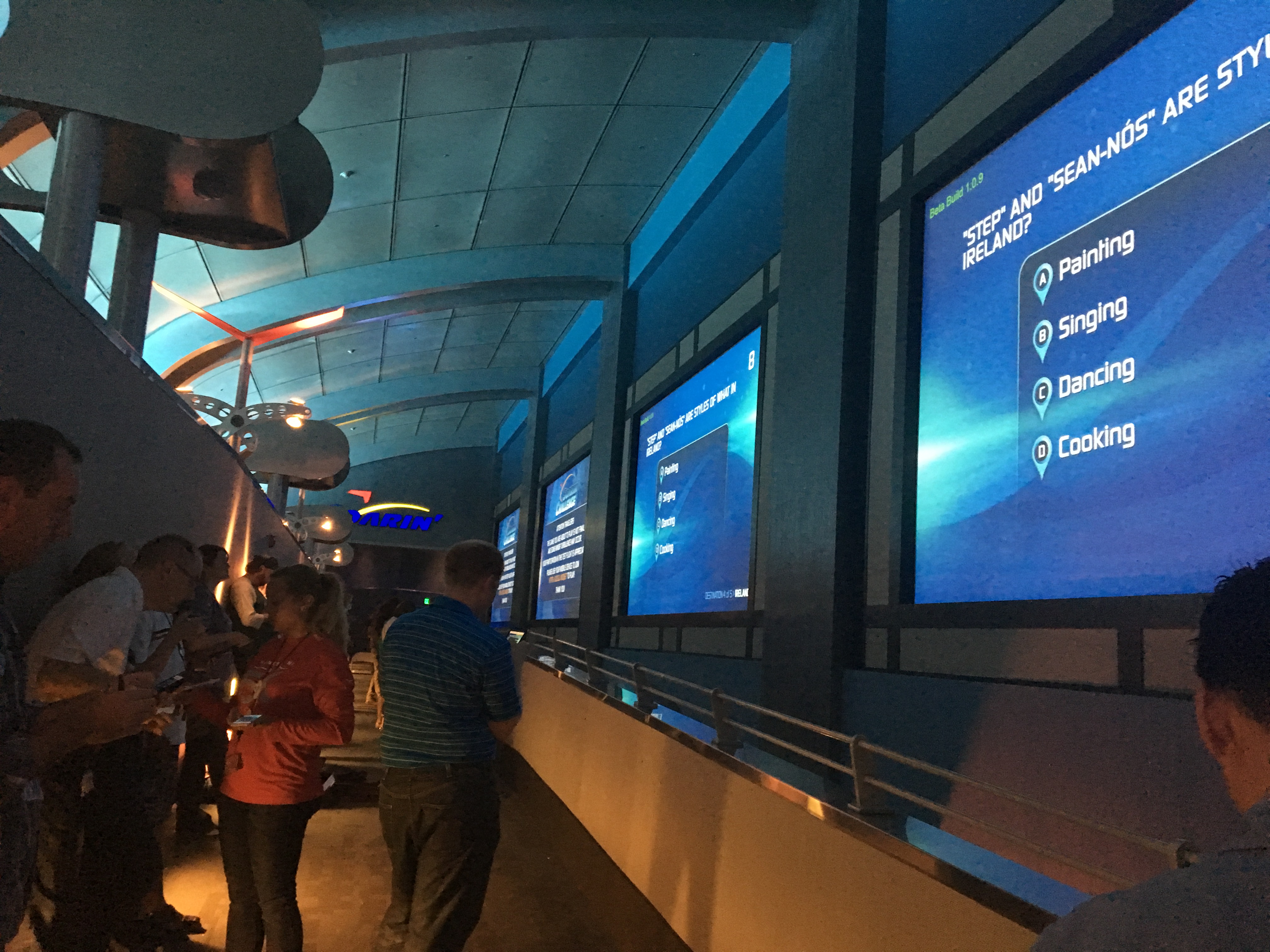 A crowd of people on their mobile phones, looking at a large screen display on the wall.